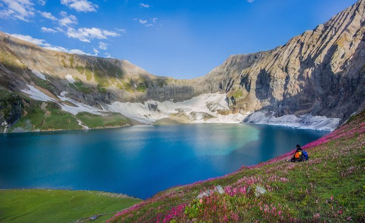 Wide photograph of Ratti Gali Lake with surrounding peaks and wildflower meadows. 