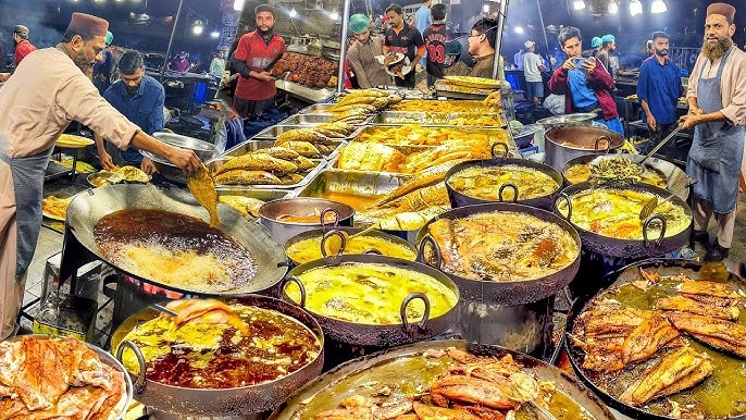 Lahori street food scene showing coal-fired karahi cooking and seekh kebabs on skewers over a grill, with smoke and the busy street behind